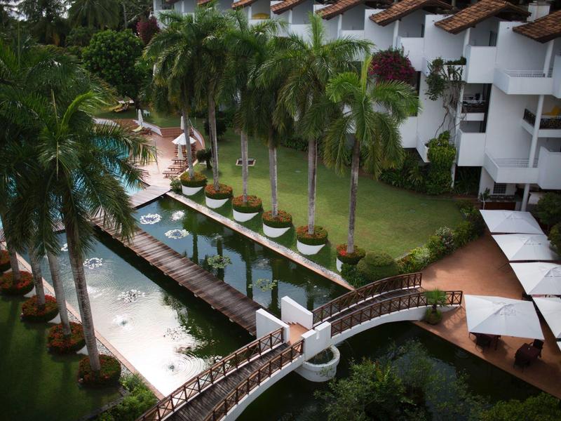 View of a tropical hotel area with a pool, bridge, and sun loungers under palm trees