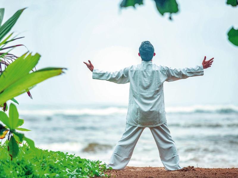 Person in white clothing stands with arms outstretched on the beach facing the sea.