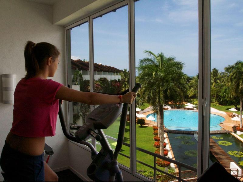 Woman exercising on a stationary bike with a view of a pool and palm trees in the hotel garden.