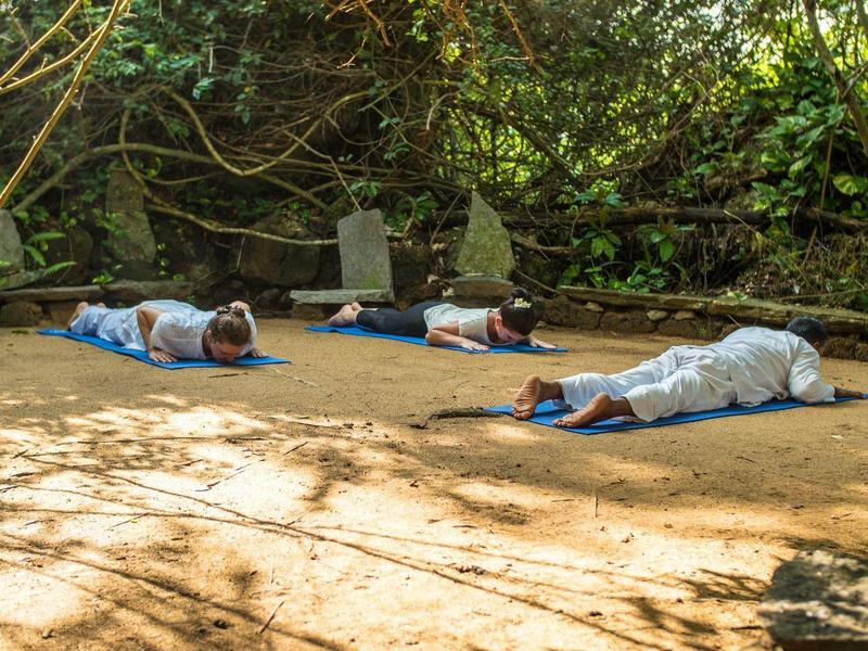 Three people lie relaxed on sandy ground in a shaded, wooded area.