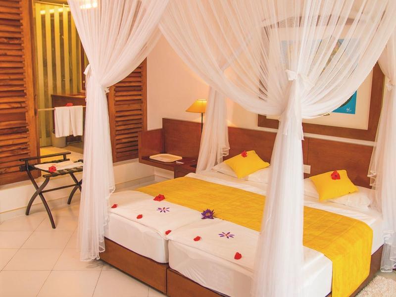 A bright hotel room with two single beds, white curtains, and yellow bedding.