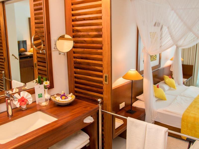 Bright bathroom with wooden furniture, sink, and view into a bedroom with double bed.
