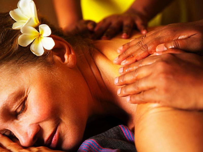 Woman with flower in hair receiving relaxing shoulder massage in warm lighting.