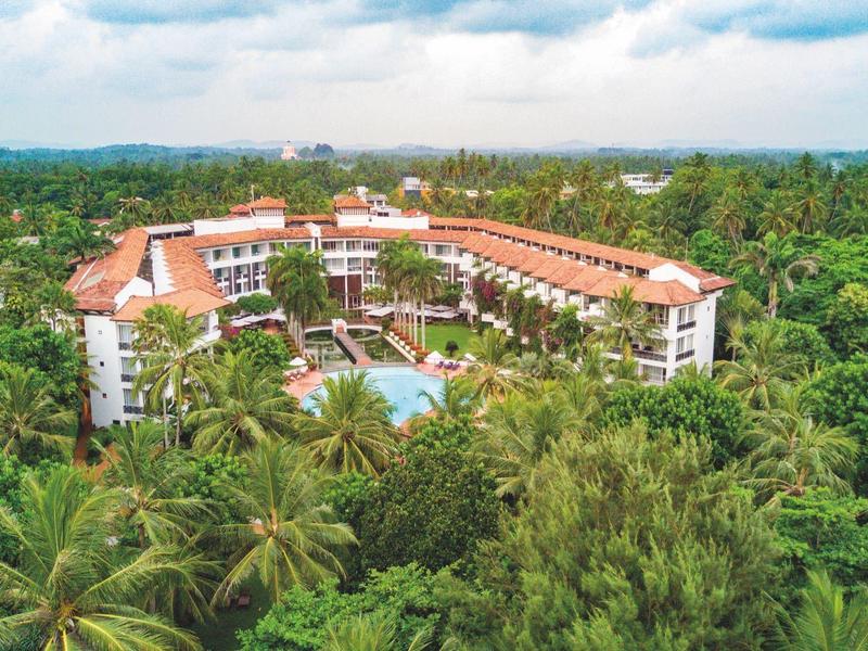 Large hotel with pool surrounded by dense green tropical forest under blue sky.