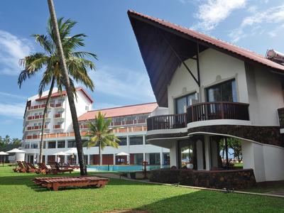 Hotelový komplex s vícepatrovou budovou a tradičně navrženou vilou, obklopený palmami a trávníkem.