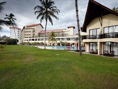 Großes Hotel mit Rasen, Pool und Palmen unter bewölktem Himmel in tropischer Umgebung.