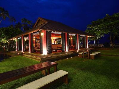 Beleuchtetes, offenes Gebäude in tropischer Umgebung bei Nacht mit Bänken und gepflegtem Rasen.