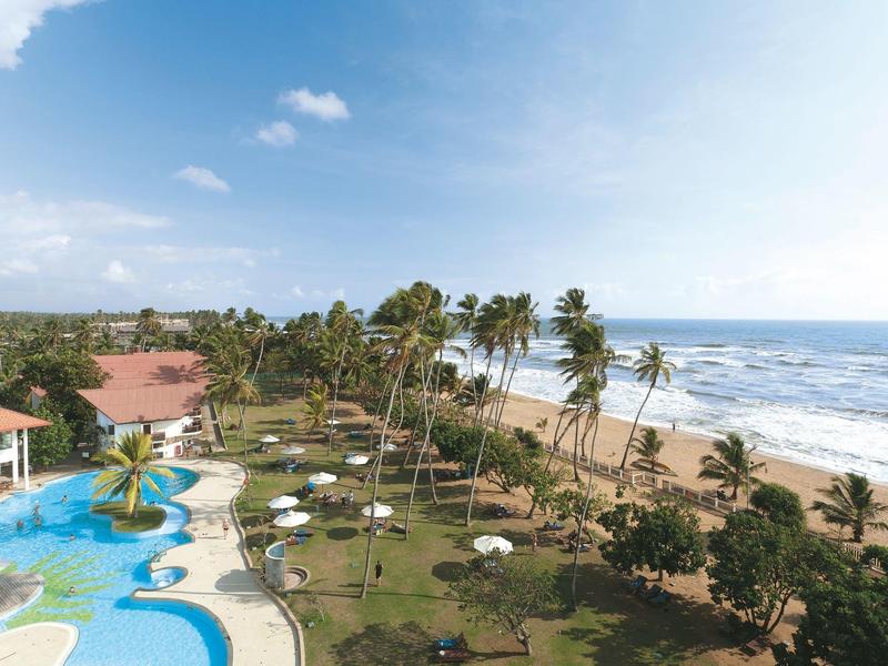 Hotelový komplex s bazény a palmami podél dlouhé písečné pláže s výhledem na moře.