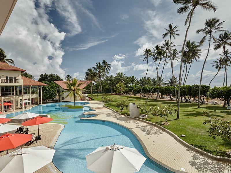 Hotelový bazén s parasoly a palmami pod oblačnou oblohou v tropickém prostředí.