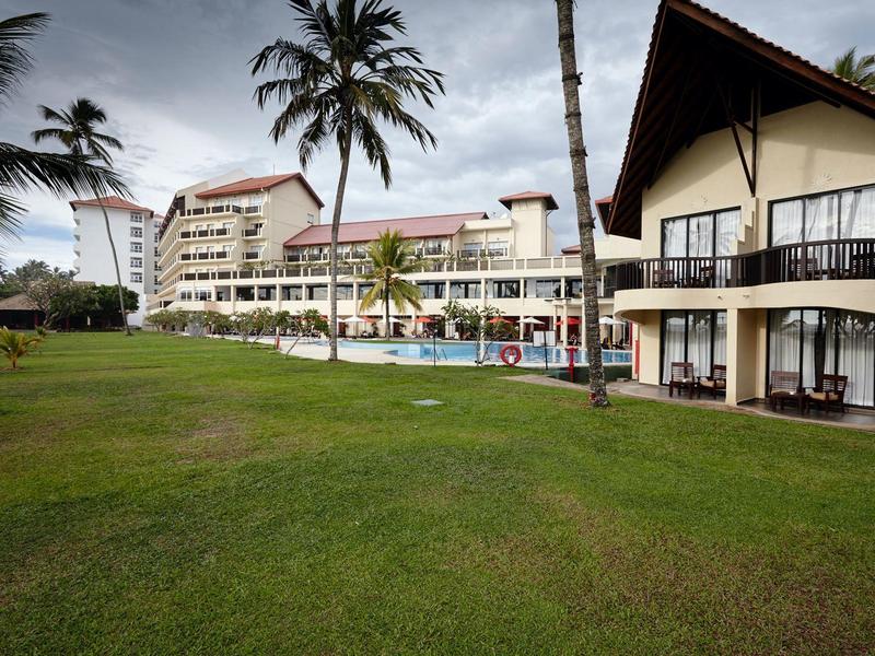 Budova hotelu s balkónovými pokoji a bazénem pod zataženou oblohou, obklopena palmami a travnatými plochami.