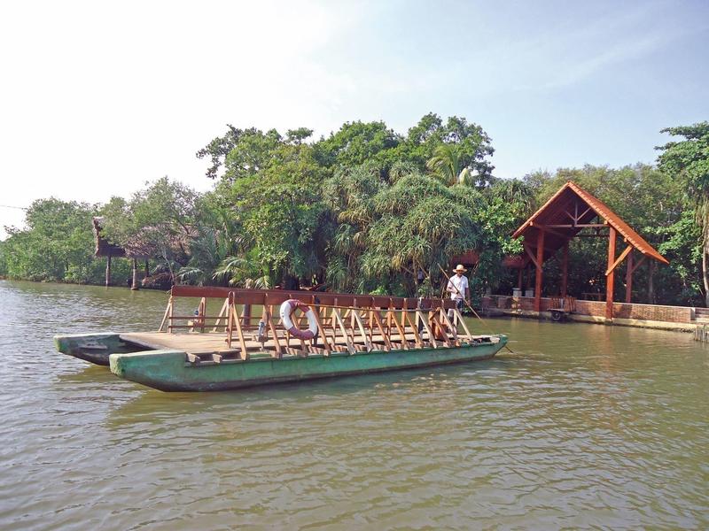 Een boot met passagiers op een rivier voor een hotel met houten gebouwen en veel groen.