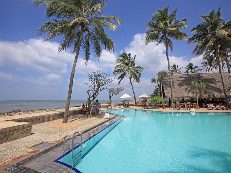 Hotelzwembad met uitzicht op het strand, palmbomen en een blauwe lucht.