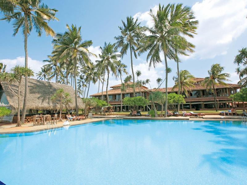 Groot hotelzwembad met palmbomen en zithoek onder blauwe lucht.