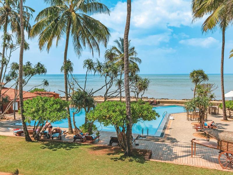 Uitzicht op een zwembad met palmbomen en de zee op de achtergrond bij zonnig weer.