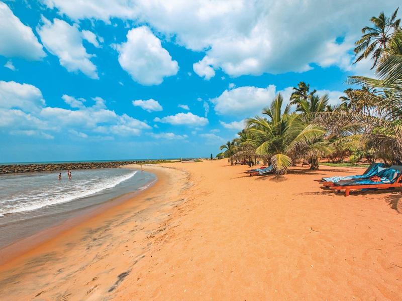 Breed zandstrand met palmbomen, ligstoelen en een blauwe lucht met wolken.