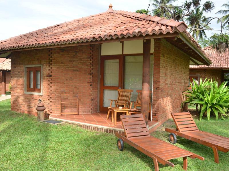 Kleine bakstenen vakantiewoning met terras, stoelen en ligstoelen op een groen gazon.