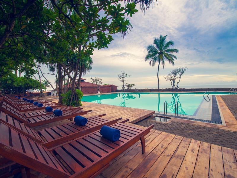 Hotelzwembad met ligstoelen en palmbomen onder bewolkte lucht bij water.