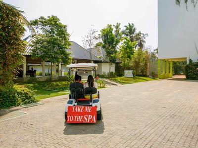 Dos personas sentadas en un carrito de golf con un letrero ‚Take me to the spa‘ frente a un jardín del hotel.