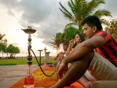 Dos personas se relajan en la playa con shisha y palmeras de fondo.