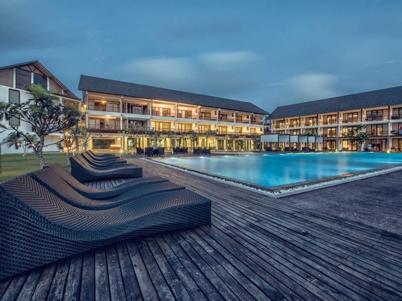 Hotel met verlicht zwembad en ligstoelen op houten terras in de avond.