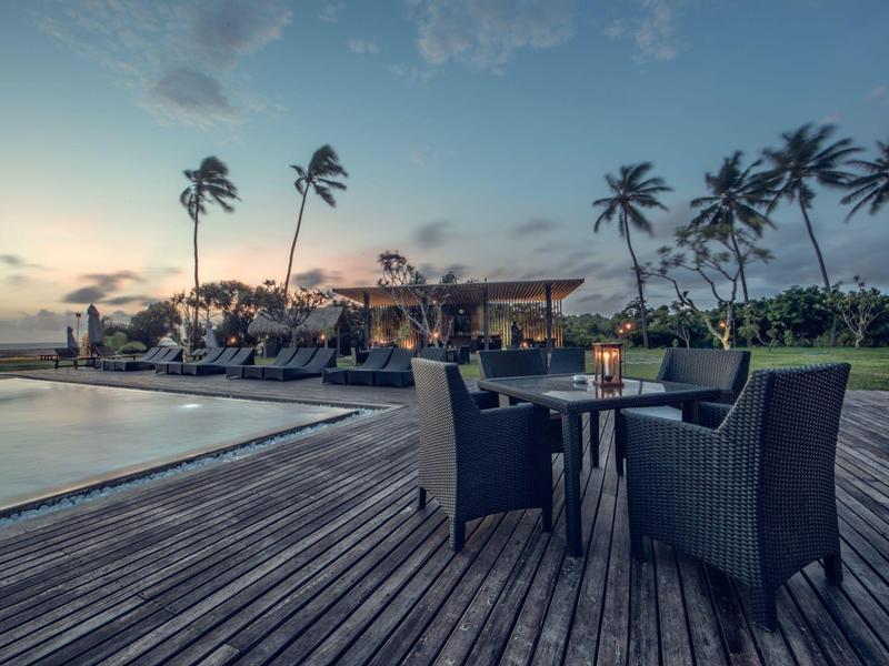 Avondterras bij het zwembad met stoelen en tafels omgeven door palmbomen en een rustige sfeer.