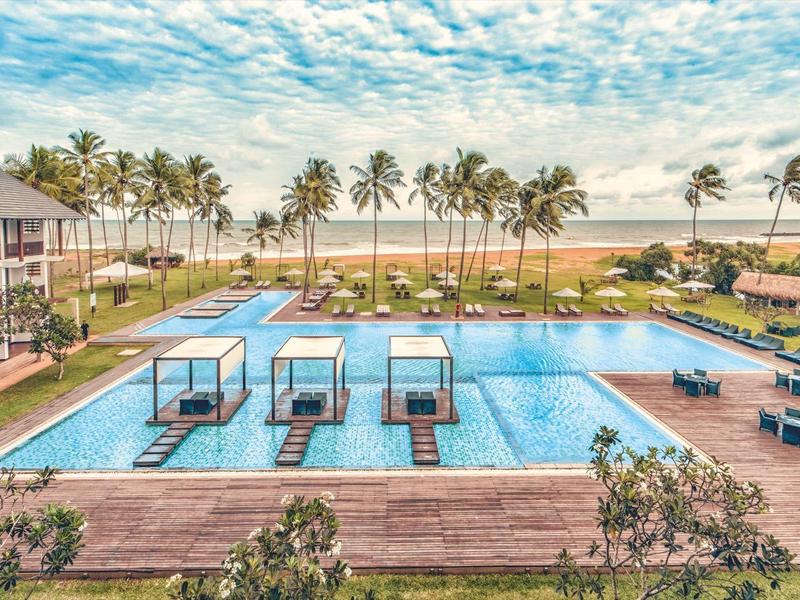 Groot hotelzwembad met overdekte ligzones en zeezicht onder bewolkte lucht.