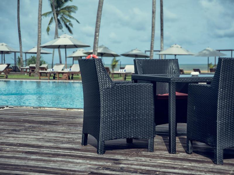 Gezellig buiten wickermeubilair op houten terras met zwembad en palmbomen op de achtergrond.