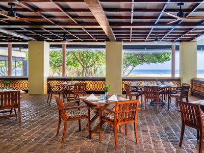 Offener Restaurantbereich mit Holztischen und Stühlen, Blick auf Meer und grüne Bäume.