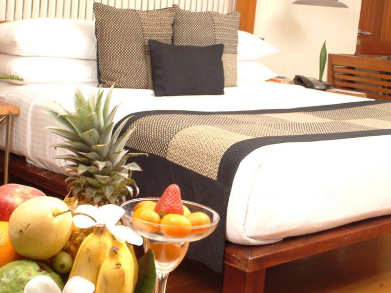 Helles Schlafzimmer mit großem Bett, weißen und grauen Kissen, Holzmöbeln und Obstschale auf Tisch.