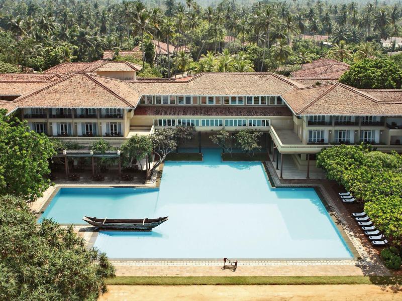 Großer rechteckiger Pool mit Steg vor Hotel mit roten Dächern, umgeben von dichtem Grün.
