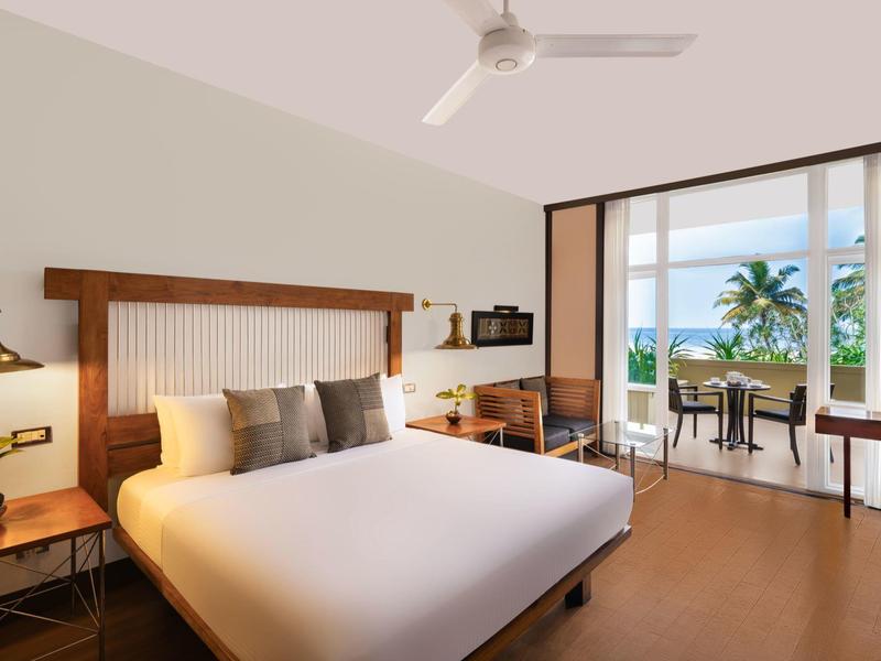 Modernes Hotelzimmer mit großem Bett, Ventilator und Blick auf Balkon mit Palme.