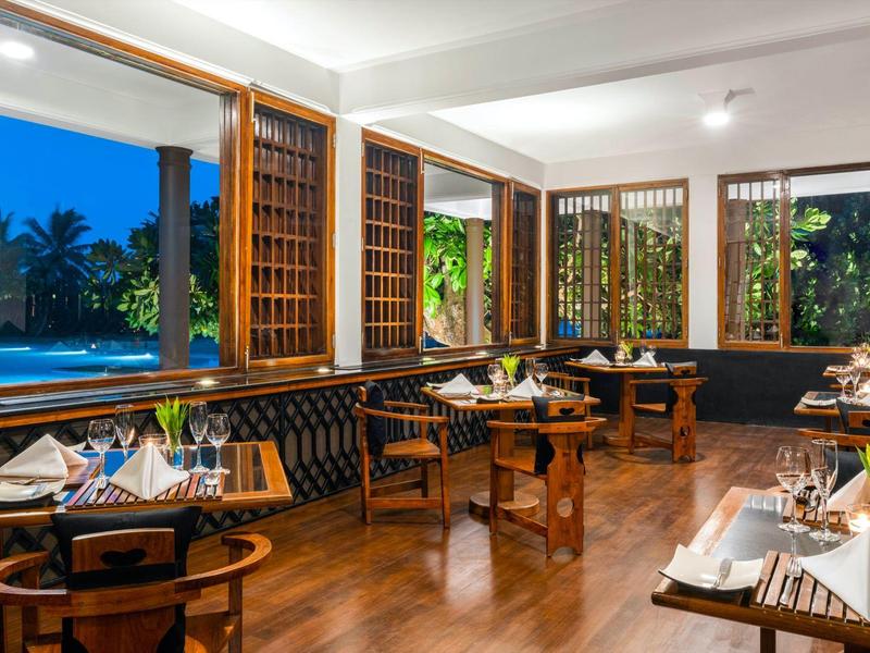 Modernes Restaurant mit Holzboden, großen Fenstern, grün dekorierten Tischen und Blick auf Pool und Palmen.
