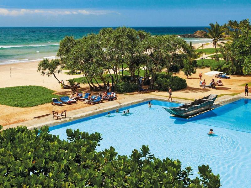 Ein großer blauer Pool mit Menschen, umgeben von grünen Bäumen und einem Strand mit Sand und Meer.