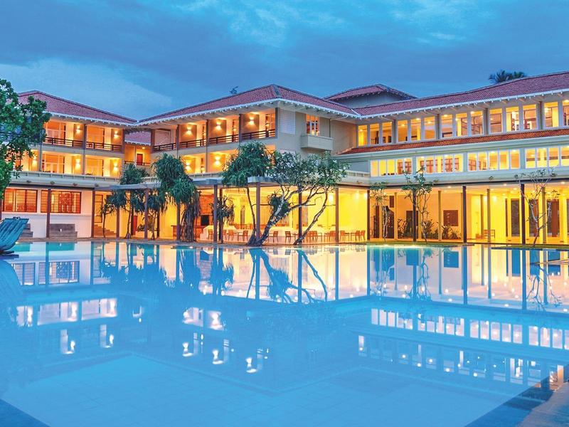 Modernes Hotelgebäude mit großen Fenstern, umgeben von Palmen und beleuchtetem Swimmingpool.