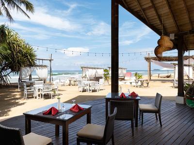 Ristorante sulla spiaggia aperto con tavoli di legno vista spiaggia sabbiosa e mare sotto cielo azzurro.