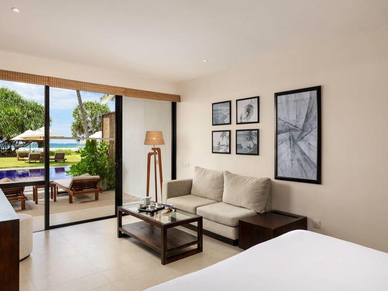 Camera d'hotel moderna con divano, tavolo in vetro e vista su terrazza con piscina e giardino.