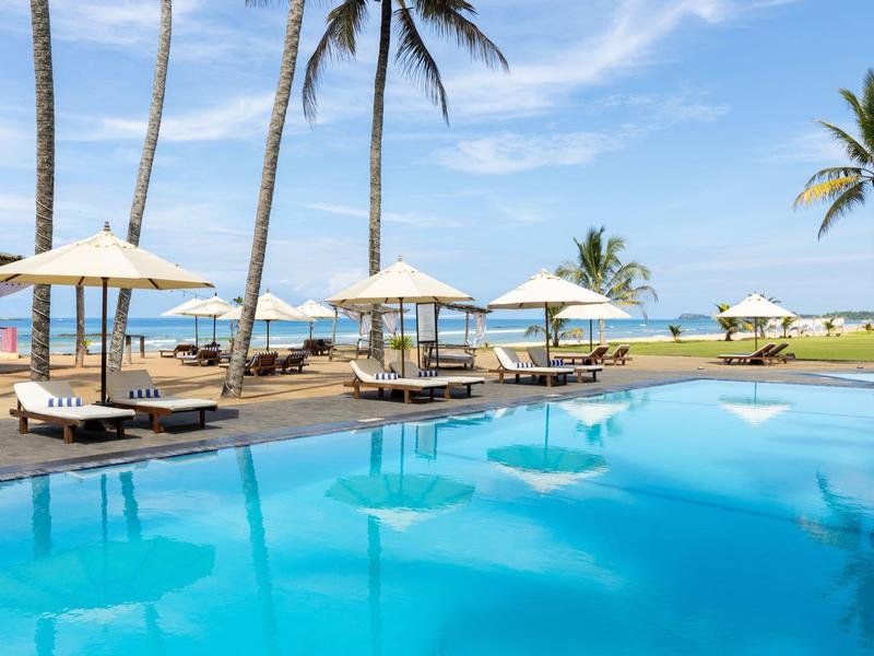 Piscina azzurra cristallina con lettini e ombrelloni davanti a palme e vista oceano.