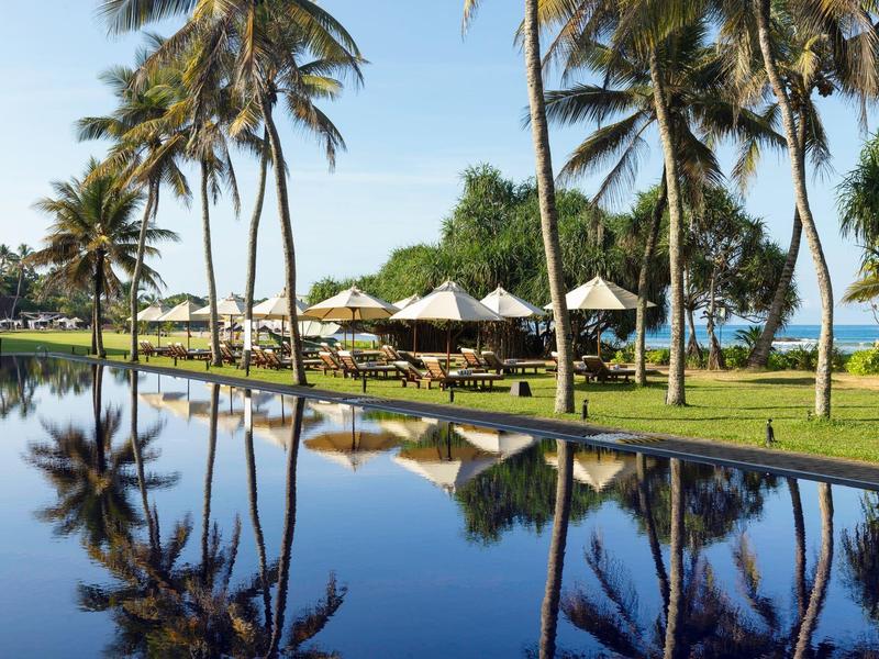 Palme affiancano una piscina tranquilla con sedie a sdraio e vista sull'oceano sotto un cielo sereno.