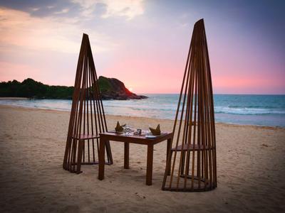 Romantyczna kolacja na plaży przy stole i unikalnych drewnianych konstrukcjach o zachodzie słońca.