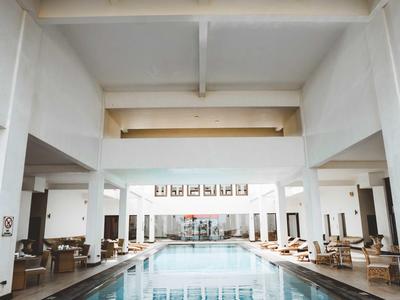 Basen kryty w jasnym hotelu z wysokimi sufitami i leżakami po bokach.