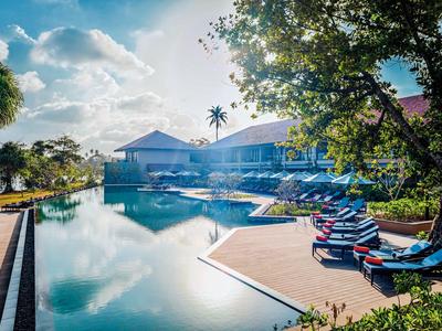 Modernes Hotel mit großem Pool und Sonnenliegen unter Bäumen am Morgen.