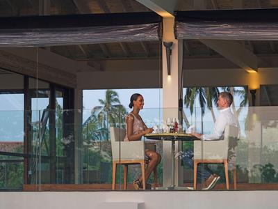 Zwei Personen sitzen beim Essen an einem Glasfenster mit Blick auf Palmen und Meer.