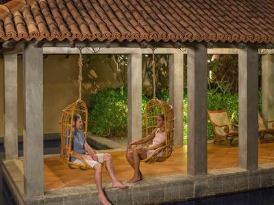 Zwei Personen entspannen abends auf Schaukelsesseln in einer überdachten Terrasse neben einem Pool.