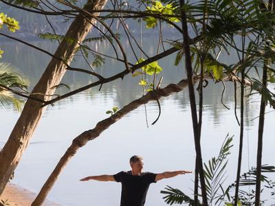 Person macht Yoga-Pose am Waldrand mit Blick auf einen ruhigen See im Hintergrund.