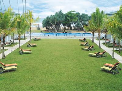 Große grüne Rasenfläche mit Liegestühlen, umgeben von Palmen und einem blauem Schwimmbecken im Hintergrund.