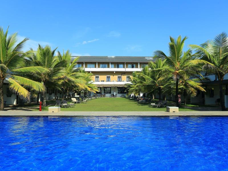 Großer blauer Pool mit Palmen und mehrstöckigem gelb-weißem Gebäude im Hintergrund.