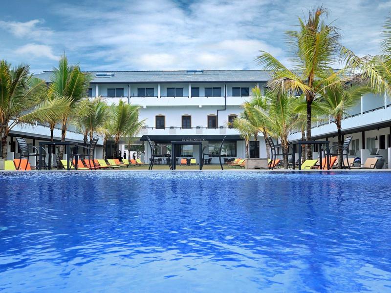 Großer blauer Pool vor einem weißen Hotelgebäude mit Palmen und Sonnenschirmen bei blauem Himmel.
