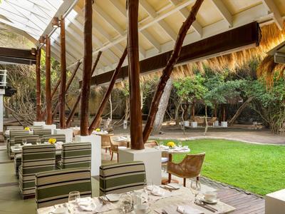 Venkovní restaurace s dřevěnou střechou, pruhovanými židlemi a výhledem na zelený trávník a stromy.