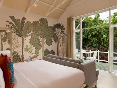 Světly hotelový pokoj s velkou postelí, pohovkami a nástěnnou malbou palem vedle terasových dveří.