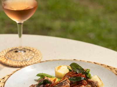 Talíř s masem v červené omáčce a zeleninou vedle sklenice rosé vína na bílém stole.